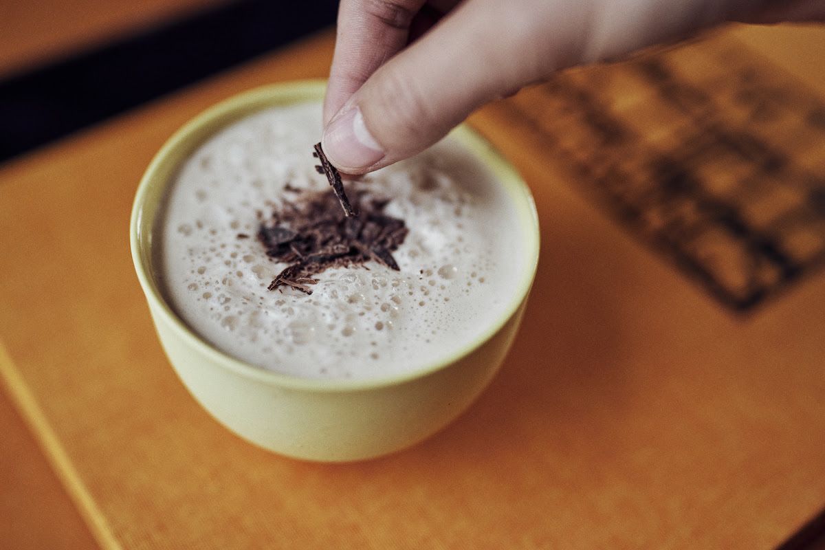 A hand places nutmeg on a foaming cup of eggnog