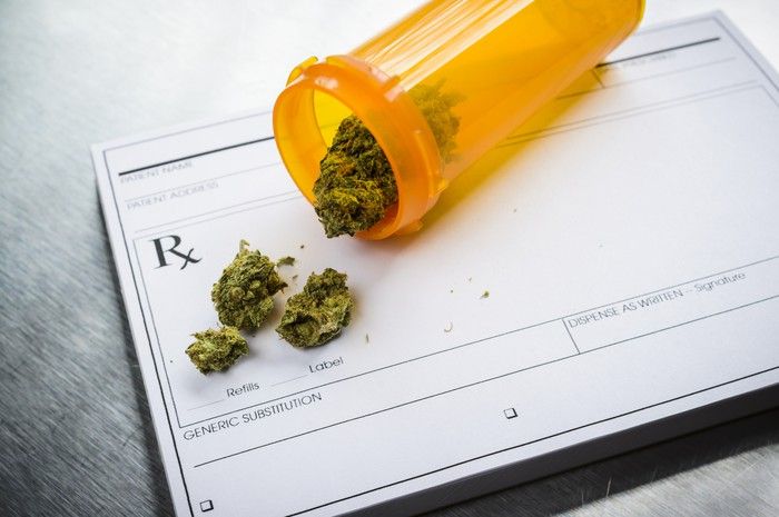 A tipped over prescription bottle containing dried cannabis buds that's lying atop a doctor's prescription pad. 