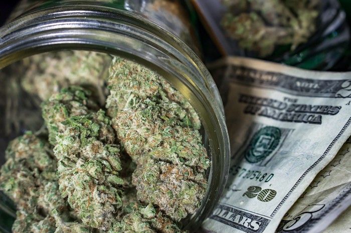 A clear jar packed with dried cannabis buds that's sitting atop a messy pile of cash.