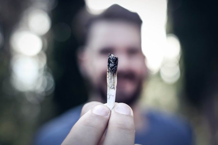 A bearded man holding a lit cannabis joint with his outstretched fingers. 