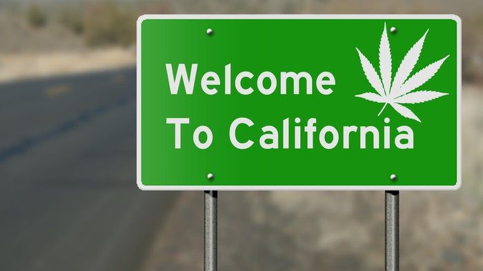 A large green highway sign that reads, Welcome to California, with a white cannabis leaf in the upper-right corner. 