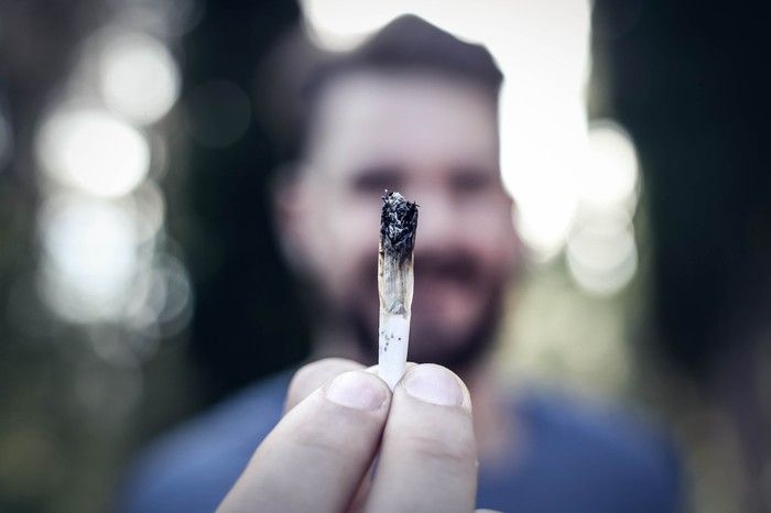A bearded man holding a lit cannabis joint with his fingertips.