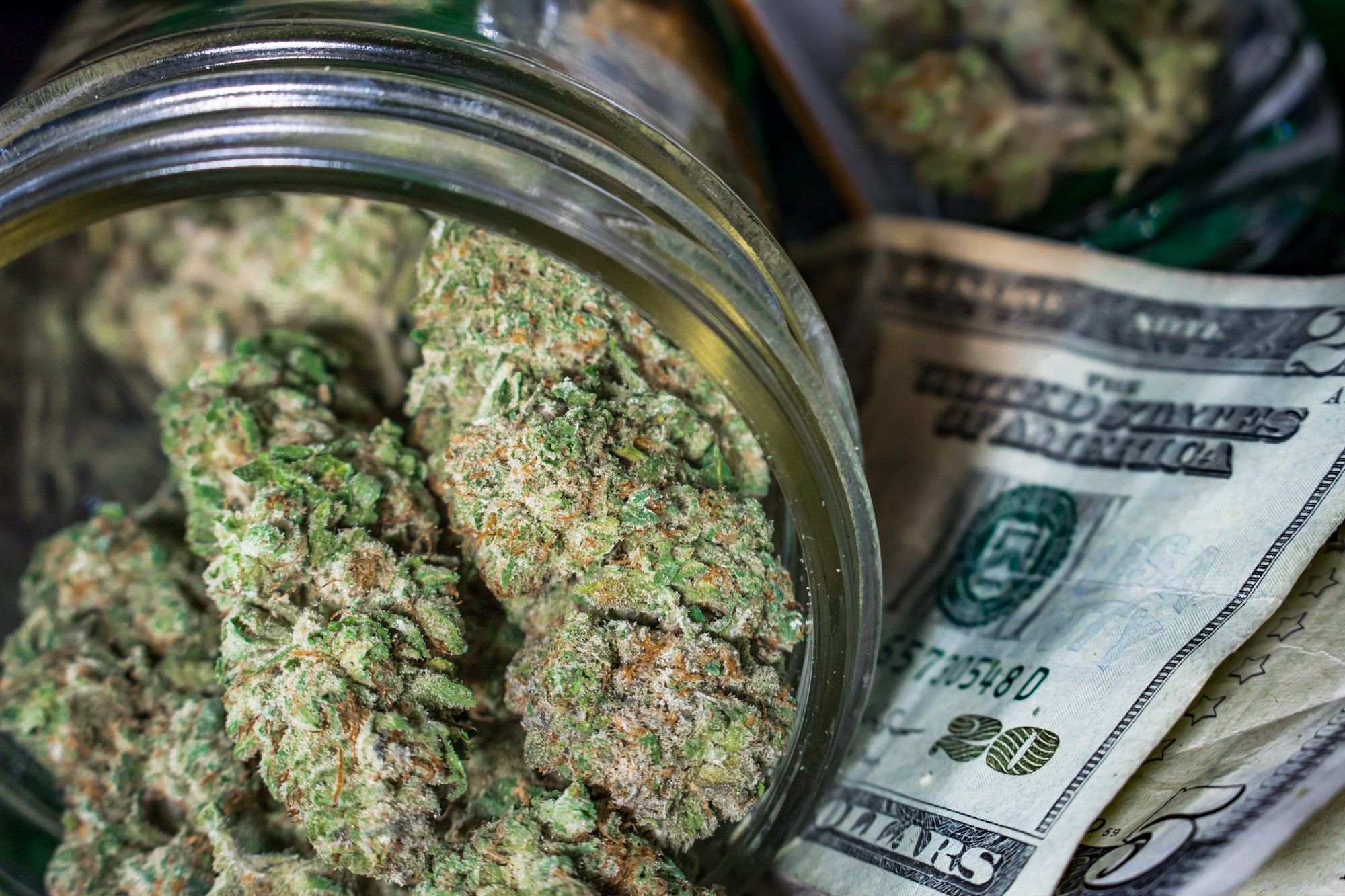 A clear jar packed with dried cannabis buds that's been tipped on its side and laid atop a small pile of cash.