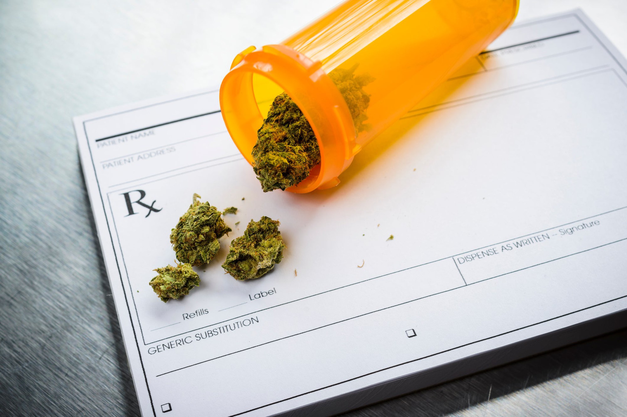 A tipped over bottle with a handful of cannabis buds inside lying atop a doctor's prescription pad.